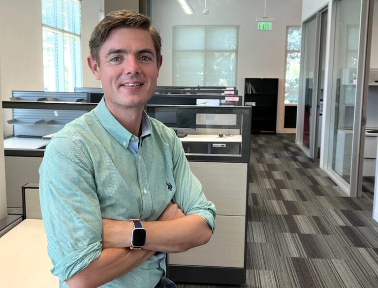 A person in a green shirt with crossed arms stands in a modern office with cubicles and large windows.