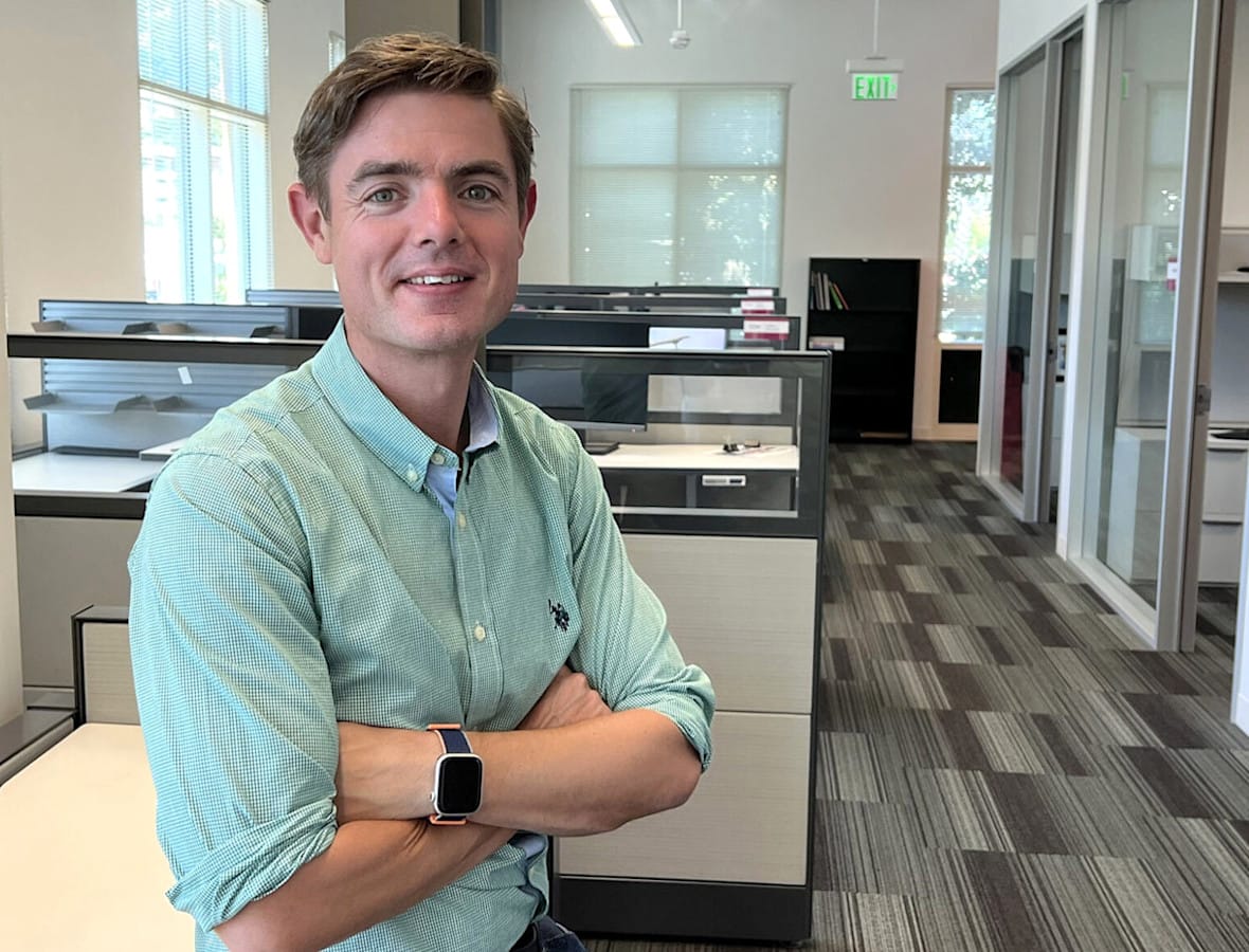 A person in a green shirt with crossed arms stands in a modern office with cubicles and large windows. A person in a green shirt with crossed arms stands in a modern office with cubicles and large windows.