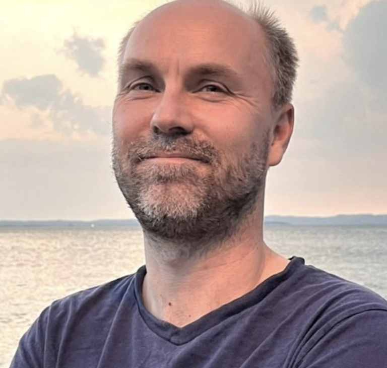 The image shows a man outdoors with a serene body of water in the background. He is smiling slightly and looking directly at the camera. The sky behind him is partially cloudy with a soft, warm sunset glow, casting gentle reflections on the water. He is wearing a plain, dark blue t-shirt and has his arms crossed. The overall ambiance of the image is calm and peaceful.