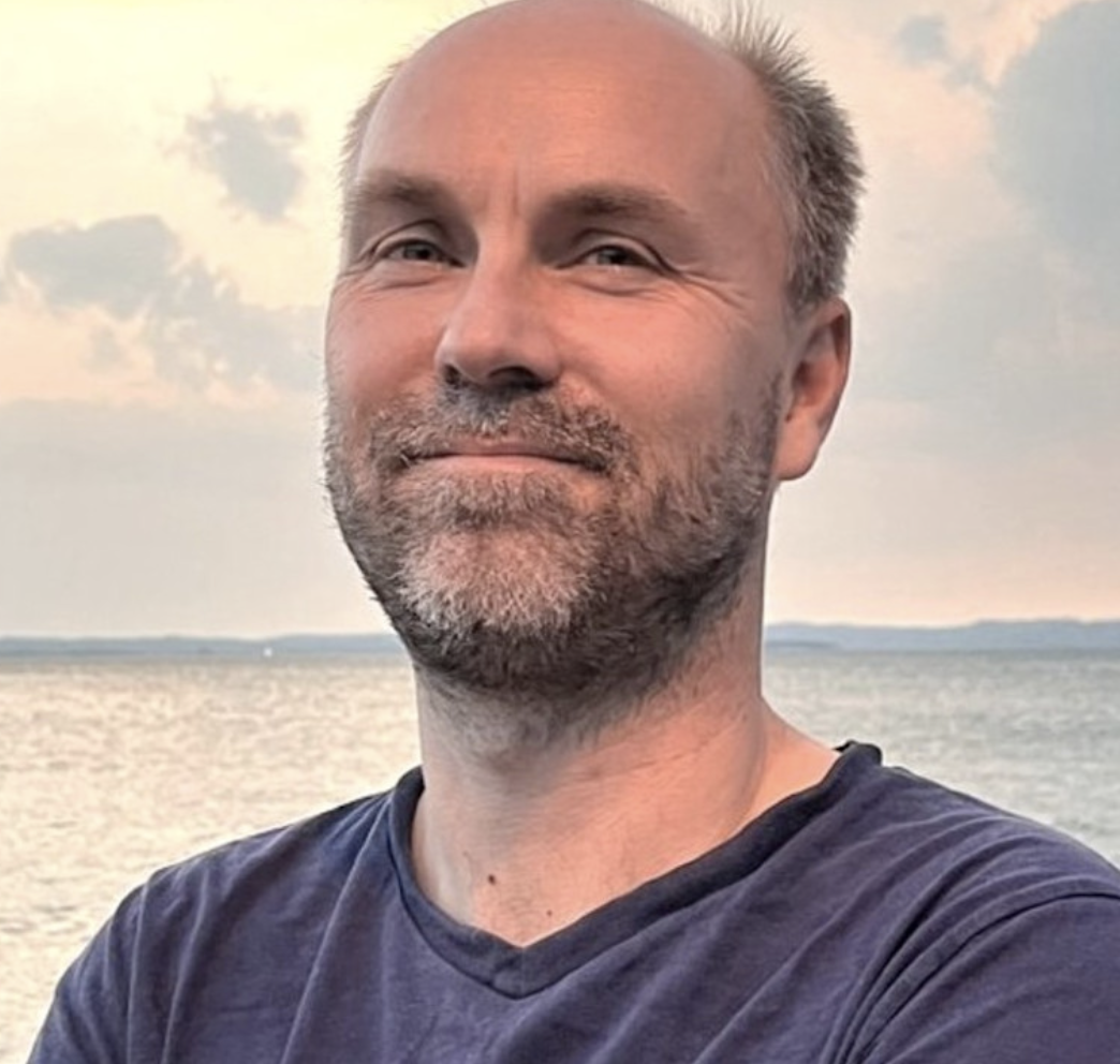 The image shows a man outdoors with a serene body of water in the background. He is smiling slightly and looking directly at the camera. The sky behind him is partially cloudy with a soft, warm sunset glow, casting gentle reflections on the water. He is wearing a plain, dark blue t-shirt and has his arms crossed. The overall ambiance of the image is calm and peaceful. The image shows a man outdoors with a serene body of water in the background. He is smiling slightly and looking directly at the camera. The sky behind him is partially cloudy with a soft, warm sunset glow, casting gentle reflections on the water. He is wearing a plain, dark blue t-shirt and has his arms crossed. The overall ambiance of the image is calm and peaceful.
